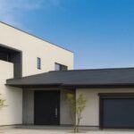 青空の下に建つ白と黒を基調としたモダンな二階建て住宅。玄関前には細い若木が2本植えられている。